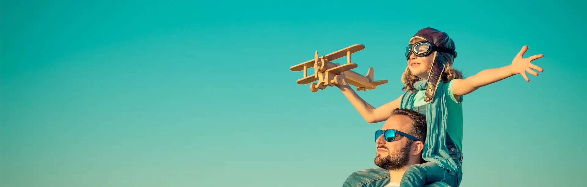 Boy with outstretched arms sitting on his father's shoulders holding out a toy wooden aeroplane